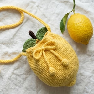 May include: A bright yellow crochet drawstring bag shaped like a lemon, featuring two green crocheted leaves and a small brown stem at the top. The bag includes a long braided yellow shoulder strap and a drawstring closure with two small yellow spheres.