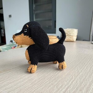 May include: A handmade crochet dachshund dog in black and tan. The dog has a long body, short legs, and floppy ears. The crochet dog is standing on a light-colored surface. The dog's body is black, and its face, ears, and paws are tan.
