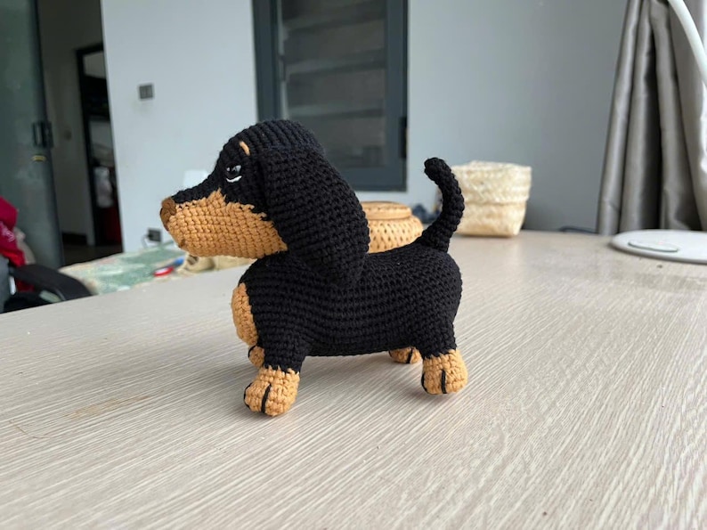 May include: A handmade crochet dachshund dog in black and tan. The dog has a long body, short legs, and floppy ears. The crochet dog is standing on a light-colored surface. The dog's body is black, and its face, ears, and paws are tan.