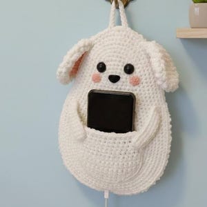 May include: A white crochet dog-shaped wall organizer with a pocket holding a black smartphone. The organizer has black button eyes, pink cheeks, and a loop for hanging. A charging cable extends from the phone.