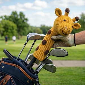 Pode incluir: Uma capa para a cabeça do taco de golfe de girafa de crochê amarela e marrom é segurada perto de uma bolsa de golfe cheia de tacos. A capa de girafa tem manchas marrons e um rosto sorridente. A bolsa de golfe é azul marinho com detalhes em couro marrom.