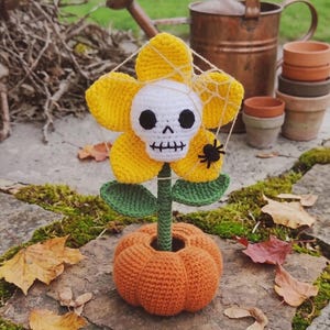 Könnte beinhalten: Eine gehäkelte gelbe Blume mit einem weißen Totenkopf in der Mitte, grünen Blättern und einem grünen Stiel, die in einer runden orangefarbenen Kürbisbasis steht. Ein dünnes weißes Spinnennetz liegt über den Blütenblättern mit einer kleinen Spinne.