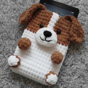 May include: A handmade crochet tablet case in the shape of a dog. The case is white with brown accents, including the ears, paws, and spots. The dog's face features black eyes and a smiling mouth. The case is designed to hold a tablet.