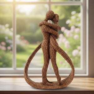 Peut inclure: Une sculpture en crochet marron représentant deux figures enlacées. Les figures sont entrelacées, les bras enroulés l'un autour de l'autre. La sculpture est posée sur une surface en bois devant une fenêtre.