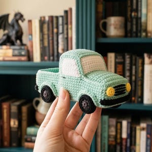 May include: A crocheted light blue pickup truck with black wheels and yellow headlights. The truck has a white cab and a detailed crochet texture. The toy truck is held in front of a bookshelf filled with books.