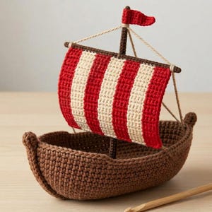 Pode incluir: Um barco de crochê artesanal com um casco marrom e uma vela listrada em vermelho e creme. O barco tem uma pequena bandeira vermelha no topo do mastro. Uma agulha de crochê de madeira está colocada perto. O barco é feito com um ponto texturizado.