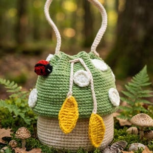 May include: A whimsical crochet mushroom-shaped bag with a cream-colored handle. The bag features a green cap with white spots, a cream-colored stem, and yellow leaf accents. A red and black ladybug is attached to the side. The bag is set against a natural, mossy background.