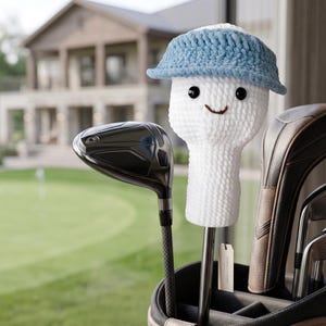 May include: A white crocheted golf club head cover featuring a light blue visor, two black bead eyes, and a small embroidered brown smile. The head cover is placed on a golf club inside a golf bag, with a small rectangular white tag attached.