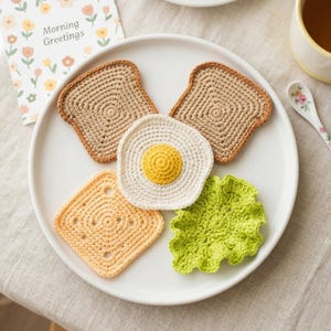 以下が含まれることがあります： 白いお皿には、かぎ針編みの食べ物が5つあります。トースト2枚、目玉焼き、正方形のトースト、レタスの葉です。トーストはタン色で、縁は茶色です。卵は黄色の黄身です。レタスは緑色です。「Morning Greetings」と書かれたカードがあります。