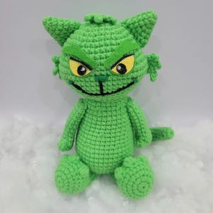 May include: A bright green crocheted plush toy shaped like a cat with pointed ears, a wide grin, and yellow felt eyes. The figure features a textured yarn surface, dark embroidered facial details, and a small tail, sitting in an upright position.