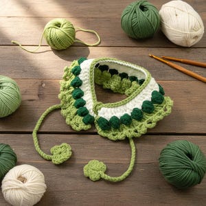 May include: A handcrafted, crocheted pet collar in white and green shades. The collar has a scalloped edge and green decorative elements. Balls of yarn in green and cream colours and crochet hooks are arranged around the collar on a wooden surface.