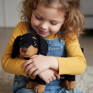 May include: A handmade crochet dachshund dog toy. The toy is black and tan, with floppy ears and a long body. The dog is being held by a person wearing a yellow long-sleeve shirt and blue denim overalls.