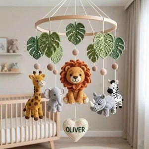 May include: A circular wooden baby mobile featuring crocheted figures of a lion, giraffe, elephant, rhinoceros, and zebra, interspersed with green crocheted monstera leaves and small spheres. A white crocheted heart at the bottom displays the name OLIVER.