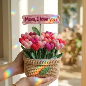 May include: A crocheted flower arrangement in a woven basket. The arrangement features heart-shaped flowers in shades of pink and red, with green leaves. A pink banner above reads "Mom, I love you."