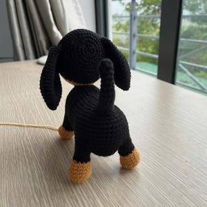 May include: A crocheted dachshund dog figurine, viewed from the rear. The dog is primarily black with tan paws and a tan underside. The ears are long and floppy. The tail is upright. The figurine is on a light-colored wooden surface.
