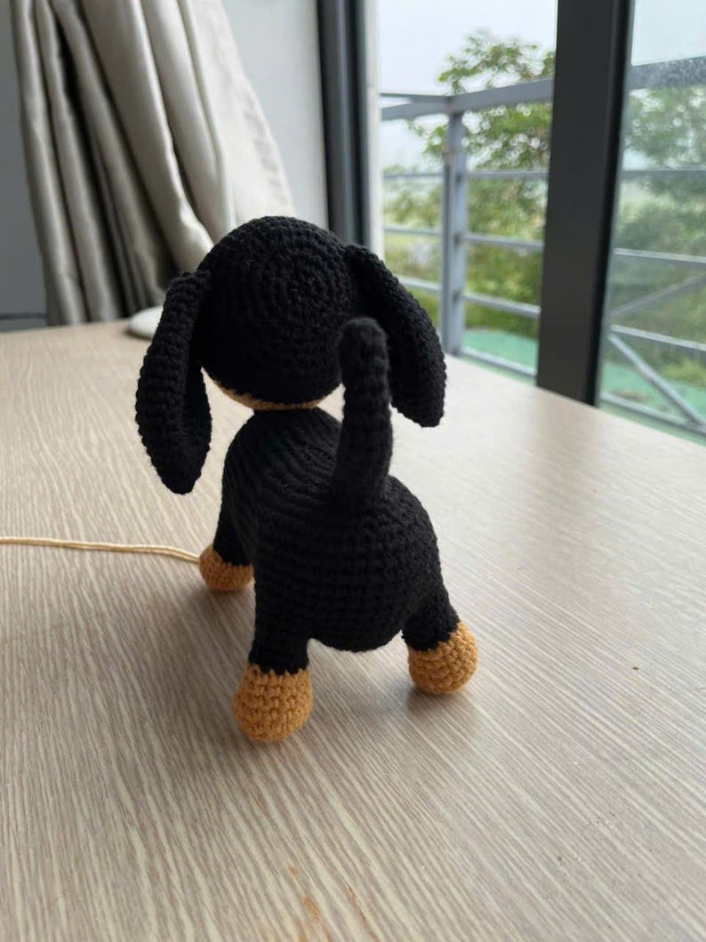 May include: A crocheted dachshund dog figurine, viewed from the rear. The dog is primarily black with tan paws and a tan underside. The ears are long and floppy. The tail is upright. The figurine is on a light-colored wooden surface.