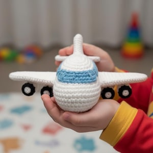 May include: A crocheted toy aeroplane in white with blue detailing on the cockpit. The plane has white wings and black wheels. The aeroplane is held in someone's hands.
