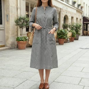 Puede incluir: Vestido midi a cuadros blancos y negros con mangas largas, cuello mandarín y cintura anudada. El vestido tiene dos bolsillos delanteros y se combina con mocasines de cuero marrón y un bolso de paja.