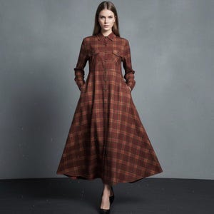 Puede incluir: Un vestido largo abotonado con estampado de cuadros marrones. El vestido tiene mangas largas, cuello camisero y dos bolsillos en el pecho. El vestido tiene una falda acampanada y se usa con tacones negros.