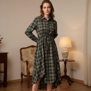 May include: A long-sleeved plaid dress in shades of green, black, and beige. The dress features a collared neckline, button-down front, and a drawstring waist. The skirt has an asymmetrical hemline. The dress is made of a soft, woven fabric.