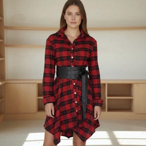May include: A red and black plaid shirt dress with long sleeves and a button-down front. The dress is cinched at the waist with a wide black belt. The dress has a collar and a slightly asymmetrical hemline.