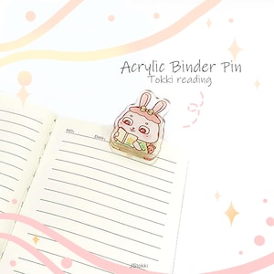 Op de afbeelding: Een acryl binder pin met een cartoon konijn dat een boek leest. De pin is transparant met roze, groene en witte accenten. De tekst "Acrylic Binder Pin Tokki reading" staat boven de pin. De pin staat op een gelijnde notitieboekpagina.
