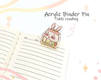 May include: An acrylic binder pin featuring a cartoon rabbit reading a book. The pin is clear with pink, green, and white accents. The text "Acrylic Binder Pin Tokki reading" is above the pin. The pin is on a lined notebook page.