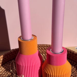 Op de afbeelding: Twee decoratieve kandelaars met roze kaarsen. De houders zijn tweekleurig, met een geribbeld ontwerp in oranje en roze. De kaarsen zijn lichtroze van kleur. De houders staan op een geweven mand.