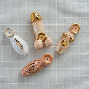 May include: Four ceramic sculptures with gold detailing. The sculptures are shaped like a penis, a vulva, and a female form. The penis sculpture is light pink with a gold tip. The vulva sculpture is white with gold detailing. The female form is light pink with gold accents.