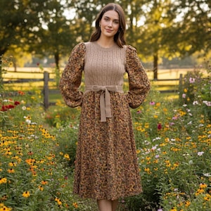 Könnte beinhalten: Ein knielanges Kleid mit einem floral bedruckten Rock und einem Zopfstrick-Oberteil. Das Kleid hat einen beigefarbenen Schleifengürtel und floral gemusterte Ärmel. Das Kleid wird im Freien in einem Blumenfeld getragen.