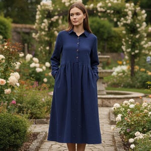 May include: A long-sleeved, navy blue dress with a collared neckline and button-down front. The dress features a gathered waist and pockets. The model is standing in a garden with white flowers and a stone pathway.