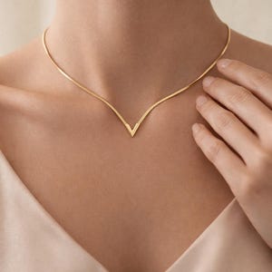 Puede incluir: Un delicado collar dorado con un colgante en forma de V. El collar es fino y se coloca cerca del cuello. El colgante tiene un diseño geométrico simple. El fondo es de un color suave y neutro.