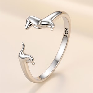 May include: A silver-colored ring featuring a dachshund dog design. The ring is open, with the dog's head and tail forming the ends. The ring is marked with "5925".