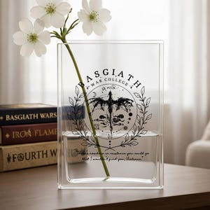 May include: A clear glass vase with a rectangular shape, featuring a black graphic of a dragon and the text "BASGIATH WAR COLLEGE." The vase holds a green stem with white flowers. Books with titles like "BASGIATH WARS" are visible.