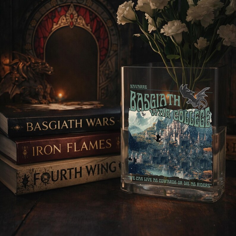 May include: A glass vase displaying a book titled "Basgiath War College" with a dragon illustration. Beside it are books labelled "Basgiath Wars", "Iron Flames", and "Fourth Wing". The vase also holds white flowers. The scene is set on a wooden surface.