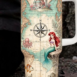 May include: White travel mug featuring a map design with a compass rose, a mermaid, and sea creatures. The mug has a white handle and lid. The map includes a building and various sea creatures like an octopus, fish, and turtle.