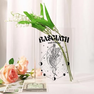 May include: A clear glass vase with the text "BASCIATH WAR COLLEGE" and a black and white illustration of a dragon wrapped around a sword. The vase holds green stems and white flowers, with pink roses in the foreground.