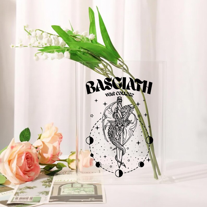 May include: A clear glass vase with the text "BASCIATH WAR COLLEGE" and a black and white illustration of a dragon wrapped around a sword. The vase holds green stems and white flowers, with pink roses in the foreground.