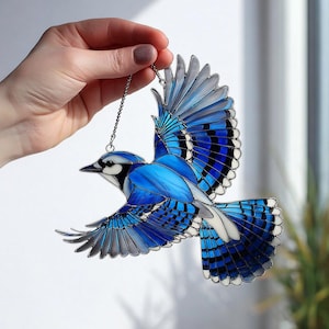 Puede incluir: Un vitral de arrendajo azul con las alas extendidas, en tonos azules, blancos y negros. El ave está suspendida por una cadena plateada, mostrando detalles intrincados. Una pieza decorativa para el hogar.