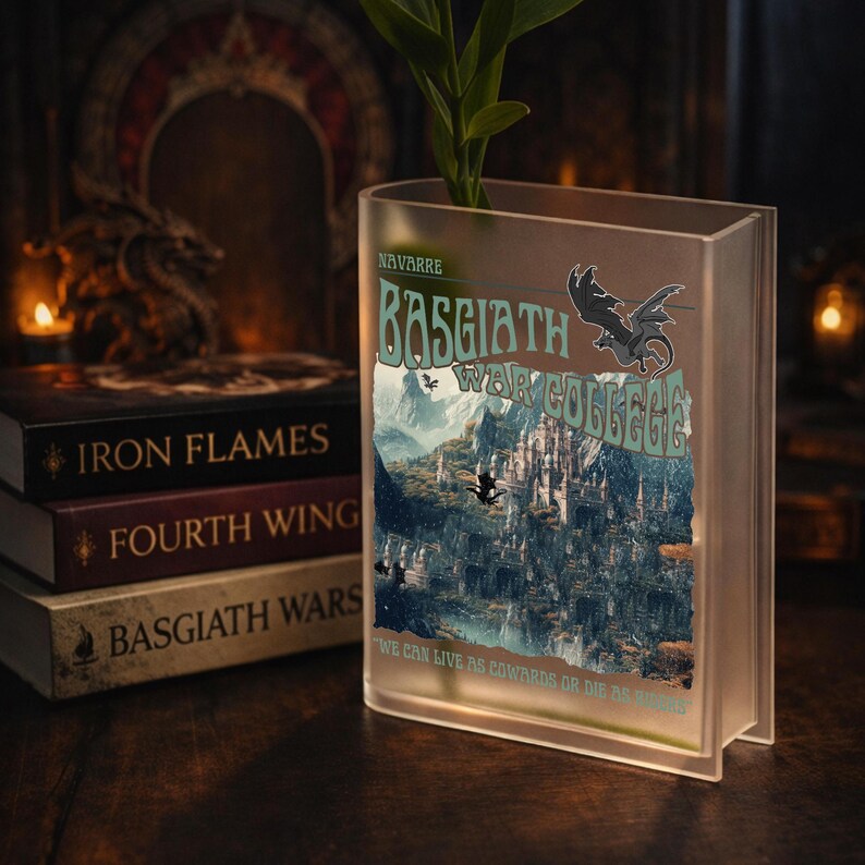 May include: A clear, book-shaped vase with the text "BASGIATH WAR COLLEGE" and a dragon illustration. The vase holds a green plant. Beside it are three books: "IRON FLAMES", "FOURTH WING", and "BASGIATH WARS". The scene is set on a dark wooden surface.