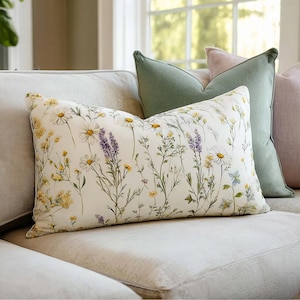 Puede incluir: Un cojín rectangular decorativo con un estampado floral de margaritas, lavanda y otras flores silvestres en amarillo, blanco y morado sobre un fondo crema. El cojín está colocado en un sofá de color claro con otros cojines verdes y rosas.