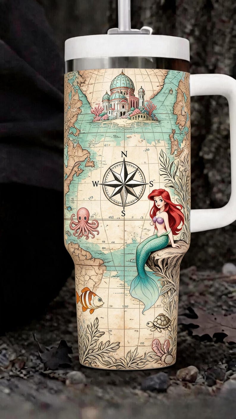 May include: White travel mug featuring a map design with a compass rose, a mermaid, and sea creatures. The mug has a white handle and lid. The map includes a building and various sea creatures like an octopus, fish, and turtle.