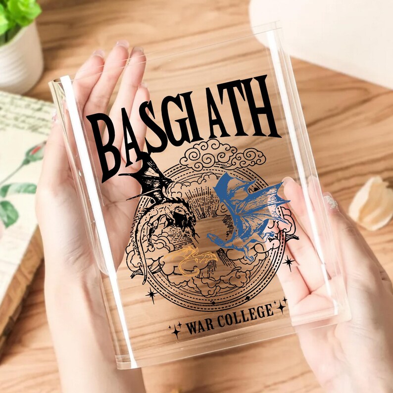 May include: Clear plastic book sleeve featuring the word "BASGIATH" in black gothic font, with a dragon illustration in black, blue, and gold. The text "WAR COLLEGE" is also visible. The sleeve is held by a person.