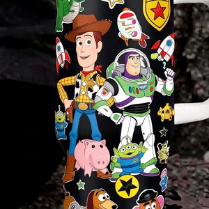 May include: A black stainless steel tumbler featuring a wrap-around graphic print of various animated characters including a cowboy, a space ranger, a green dinosaur, a pink pig, a dachshund, and a potato figure, with a white lid and handle.