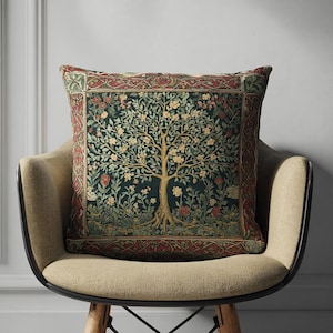 Könnte beinhalten: Quadratisches Zierkissen mit detailliertem Baumdesign. Das Kissen hat einen dunkelgrünen Hintergrund mit einem Baum in Braun-, Gelb- und Grüntönen. Der Rand ist rot mit floralen Mustern. Das Kissen liegt auf einem beigen Stuhl.
