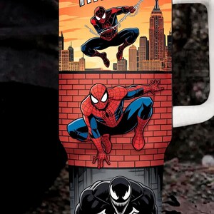 May include: A stainless steel tumbler with a white lid and handle featuring a graphic print of Spider-Man swinging over a city skyline with the text THWP!, Spider-Man crouching on a brick wall, and Venom with the text WITH GREAT POWER...