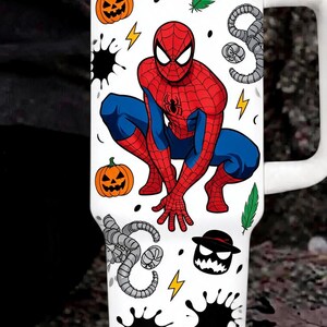 May include: A white stainless steel tumbler with a handle and lid featuring a graphic of Spider-Man in a crouching pose. The design includes orange jack-o'-lanterns, green feathers, yellow lightning bolts, black ink splatters, and gray tentacles.