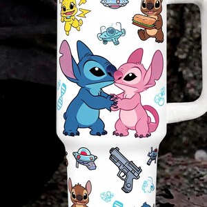 May include: A white stainless steel tumbler with a handle and lid, featuring a repeating pattern of blue and pink alien characters, a yellow creature, a grey handgun, blue DNA strands, small spaceships, and various small blue graphic icons.