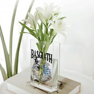 May include: A clear rectangular vase with the word "BASGLATH" in black lettering, and a graphic design. The vase holds white lilies with green stems and leaves. The vase is sitting on a white surface.