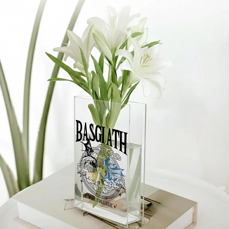 May include: A clear rectangular vase with the word "BASGLATH" in black lettering, and a graphic design. The vase holds white lilies with green stems and leaves. The vase is sitting on a white surface.